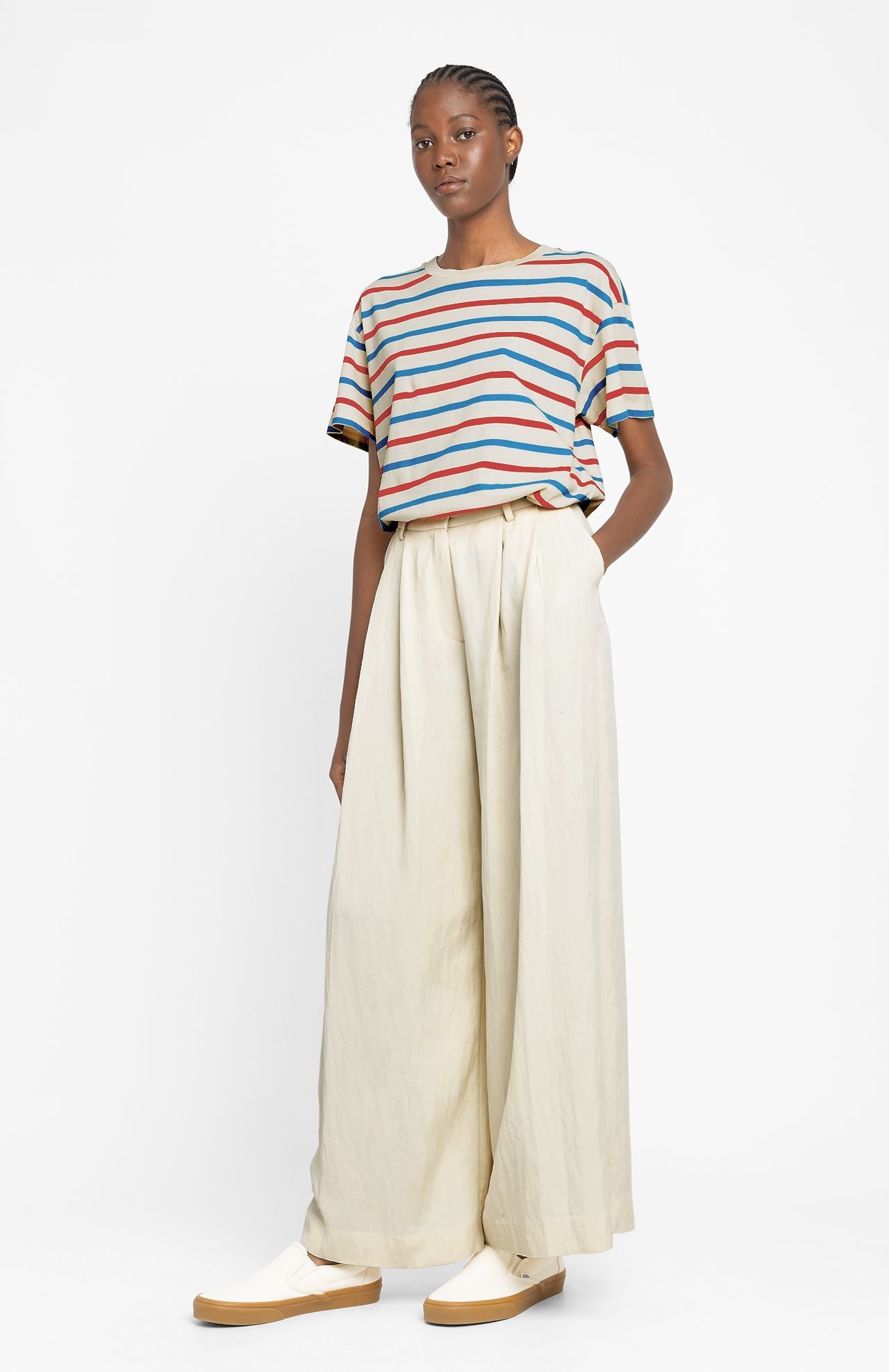 Model wears a stripe tshirt and cream pleated trousers with white trainers by Loreak Mendian