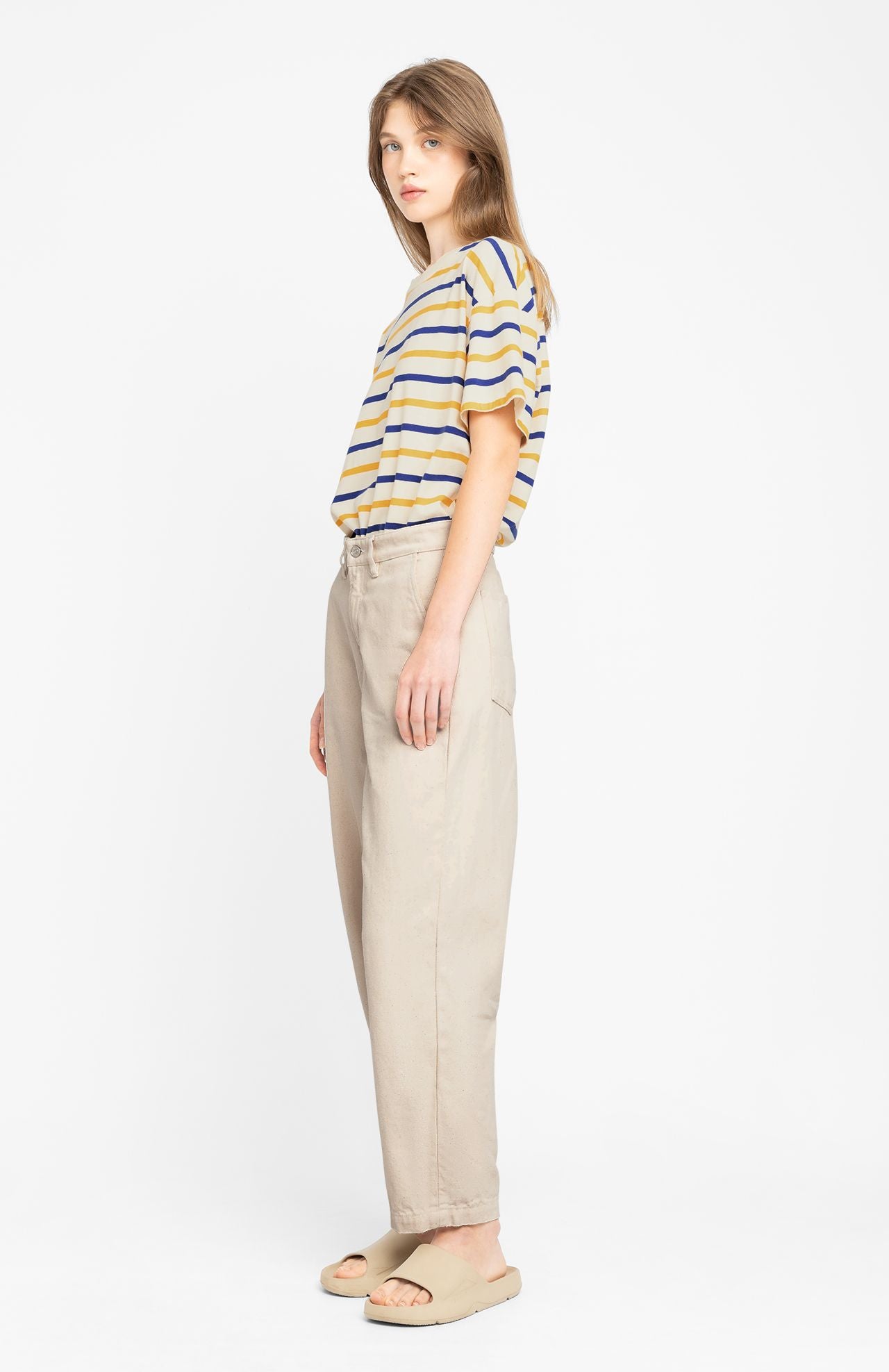 A person wearing a striped t-shirt, beige Mirena Trouser - Ecru from Loreak Mendian, and sandals stands against a white background. The outfit exudes the comfort of organic cotton jeans, ideal for those who value effortless style.