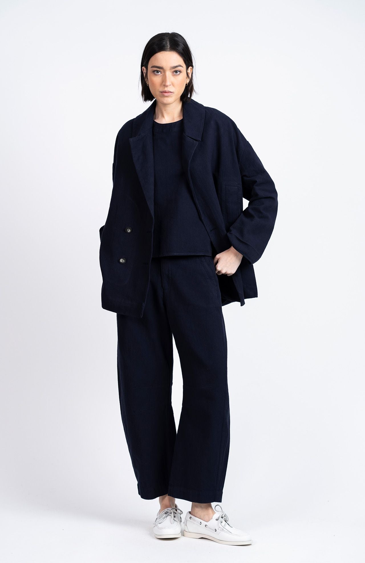 A person stands against a white background wearing Loreak Mendian's Tome Barrel Trousers in navy, paired with a matching top and white sneakers, hands in jacket pockets.