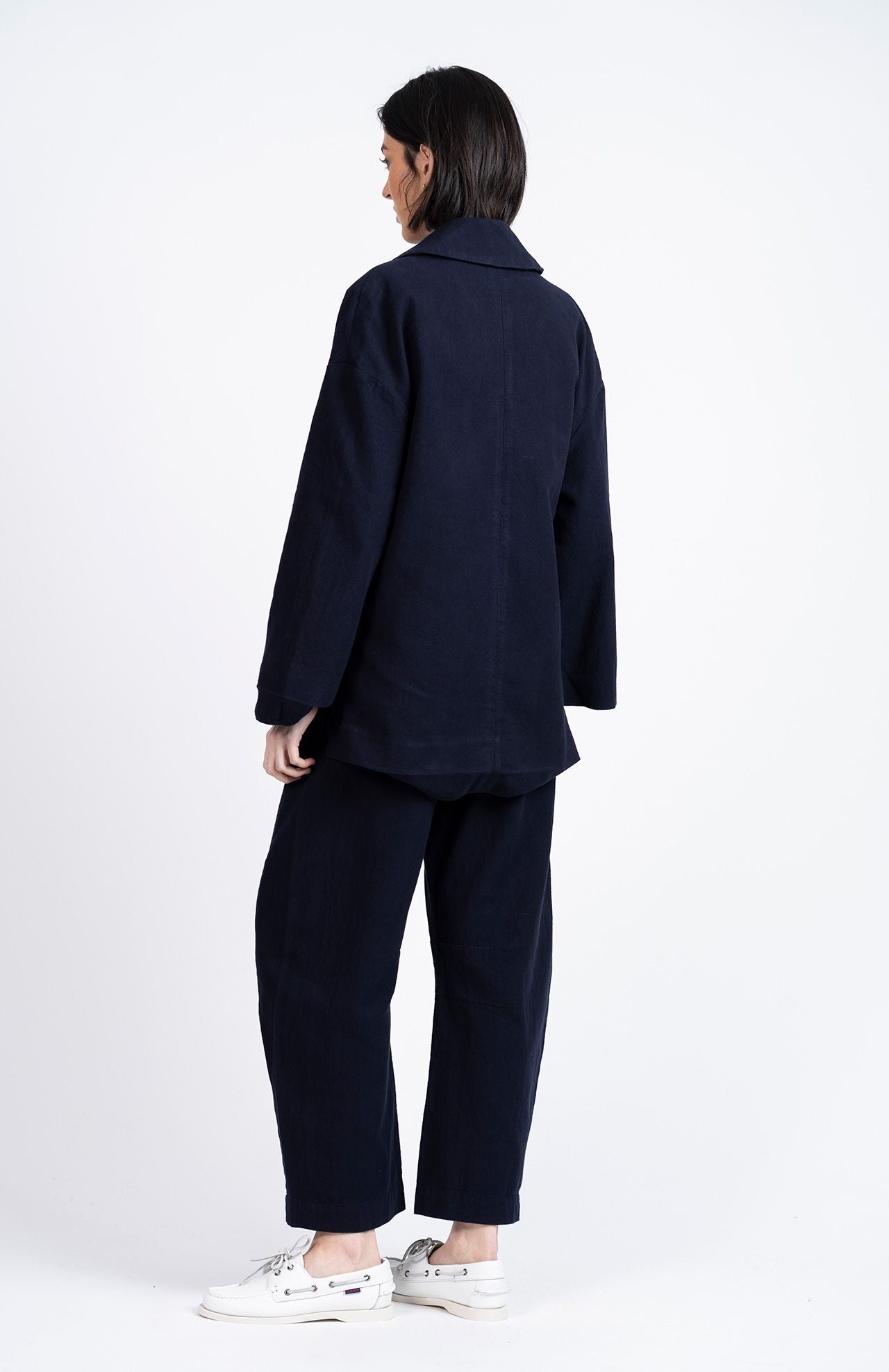 A person stands against a white background, facing away, wearing Loreak Mendian's Tome Barrel Trousers in Navy paired with a matching loose-fit jacket and white sneakers.