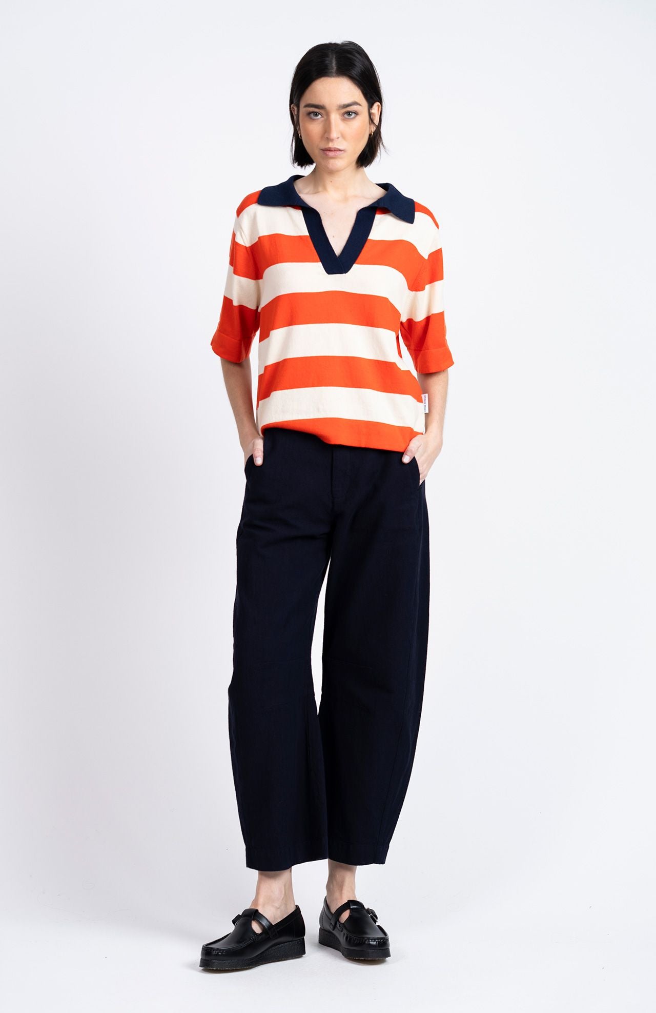 A woman stands against a white background in a red and white striped shirt with a navy collar, black loafers, and Loreak Mendian’s Tome Barrel Trousers in navy.