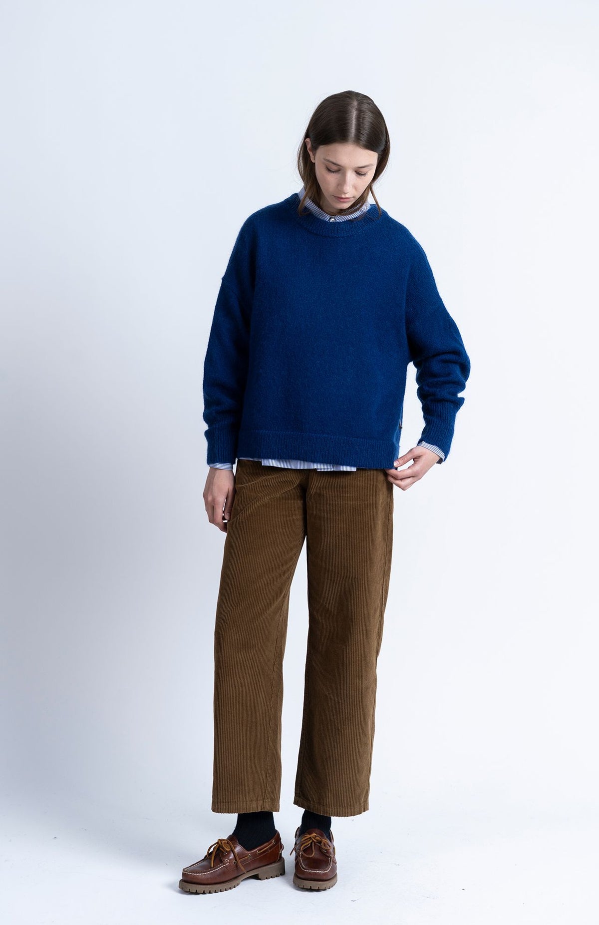 A model stands against a light gray studio background wearing a navy blue knit sweater, brown wide-leg trousers, dark socks, and brown leather loafers.