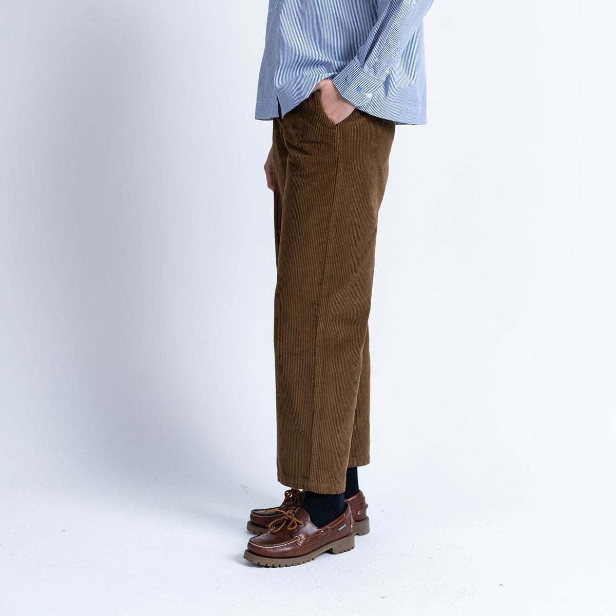A person stands sideways against a plain white background, wearing a blue shirt, black socks, brown leather shoes, and Loreak Mendian’s Bana Barrel Leg Cord Trousers in Dark Camel made from 100% cotton.