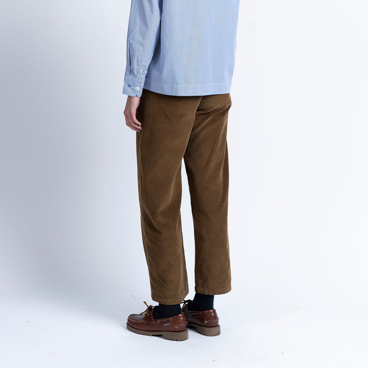 A person is shown from behind, standing against a plain white background, wearing a light blue button-down shirt, black socks, brown boat shoes, and Loreak Mendian Bana Barrel Leg Cord Trousers in Dark Camel made from 100% cotton.