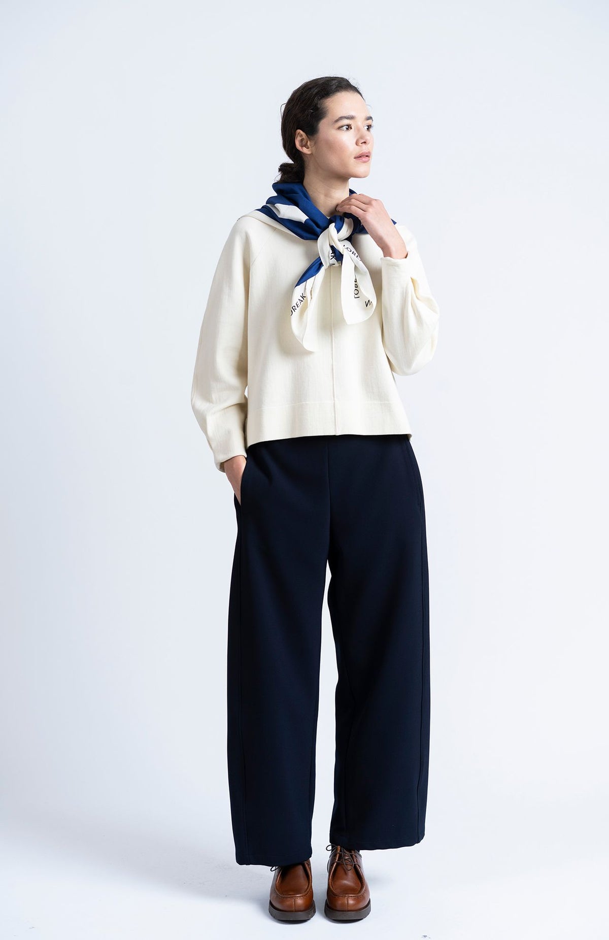 a model wears navy barrel shaped jogger trousers by loreak mendian with brown shoes and a cream knit on a white background.