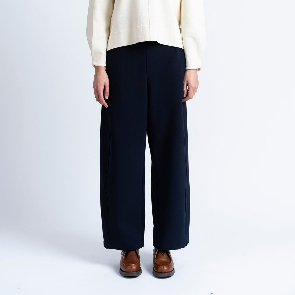a model wears navy barrel shaped jogger trousers by loreak mendian with brown shoes and a cream knit on a white background.