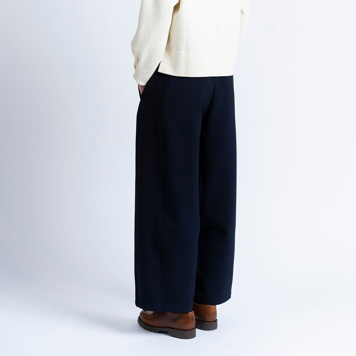 a model wears navy barrel shaped jogger trousers by loreak mendian with brown shoes and a cream knit on a white background.