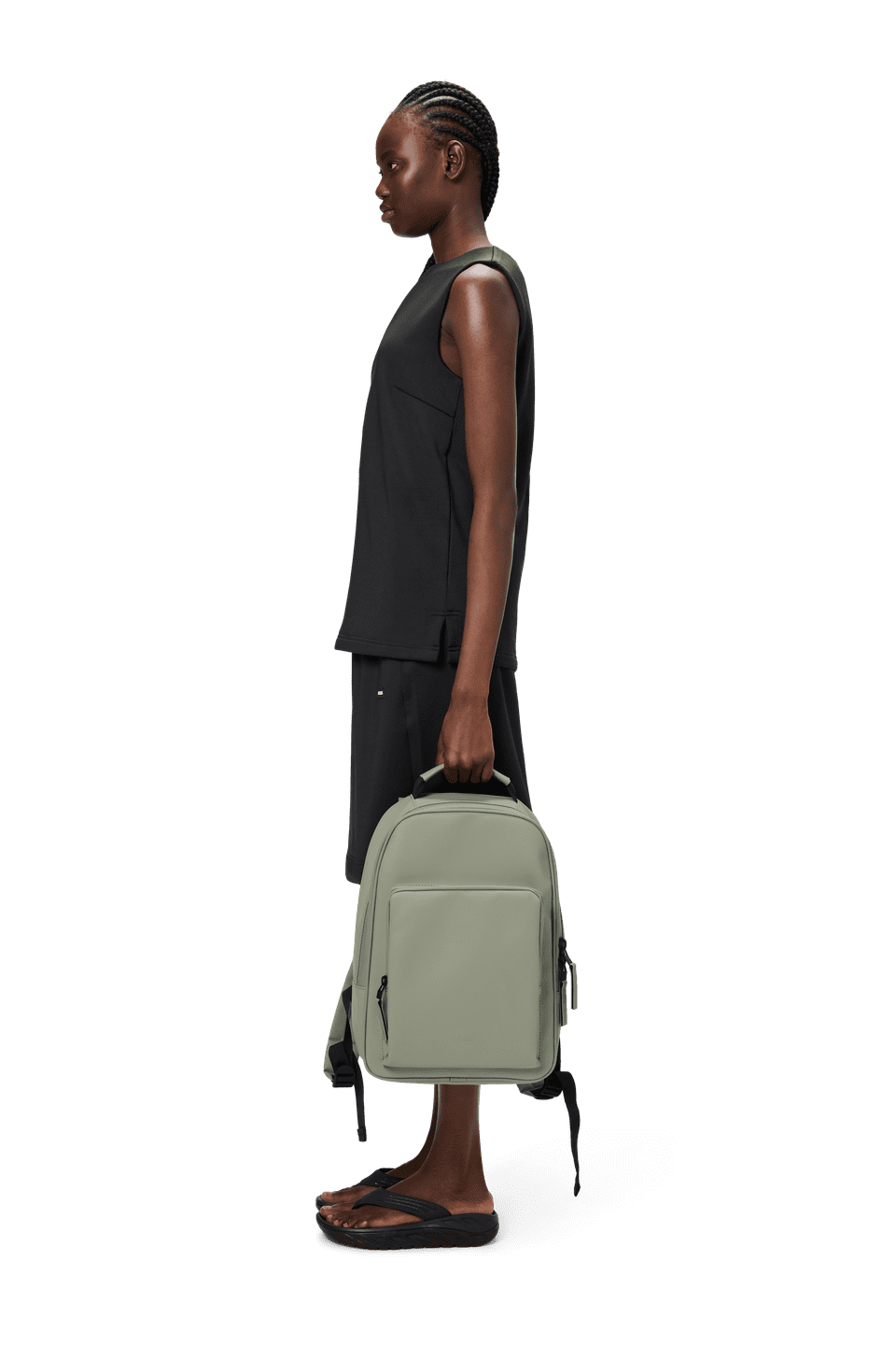 A person in a black outfit stands in profile, holding the Rains Book Daypack, which is waterproof and designed in light gray with a padded pocket for a 15" laptop.