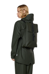 person is wearing green jacket and pants, facing the left and wearing the Green waterproof rucksack by Rains - Studio set up white backgorund 