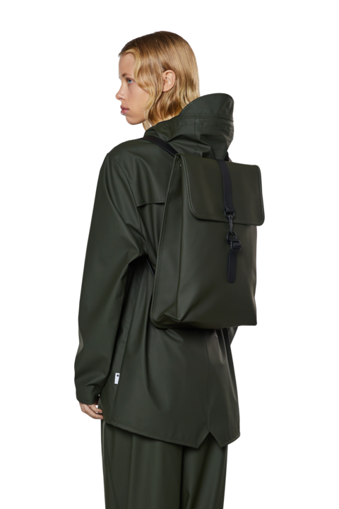 person is wearing green jacket and pants, facing the left and wearing the Green waterproof rucksack by Rains - Studio set up white backgorund 