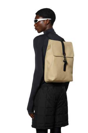 person wearing all black faces the side and is wearng the Sand waterproof rucksack by Rains - Studio set up white backgorund 