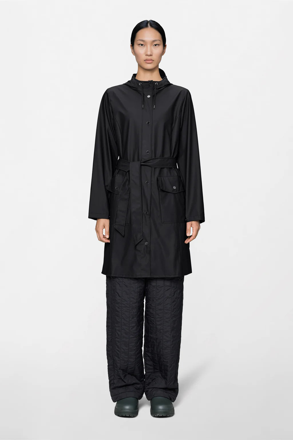 A person stands facing forward against a plain light background, wearing the Rains Curve W Jacket W3—a black belted women's raincoat—paired with quilted black pants and black waterproof shoes.