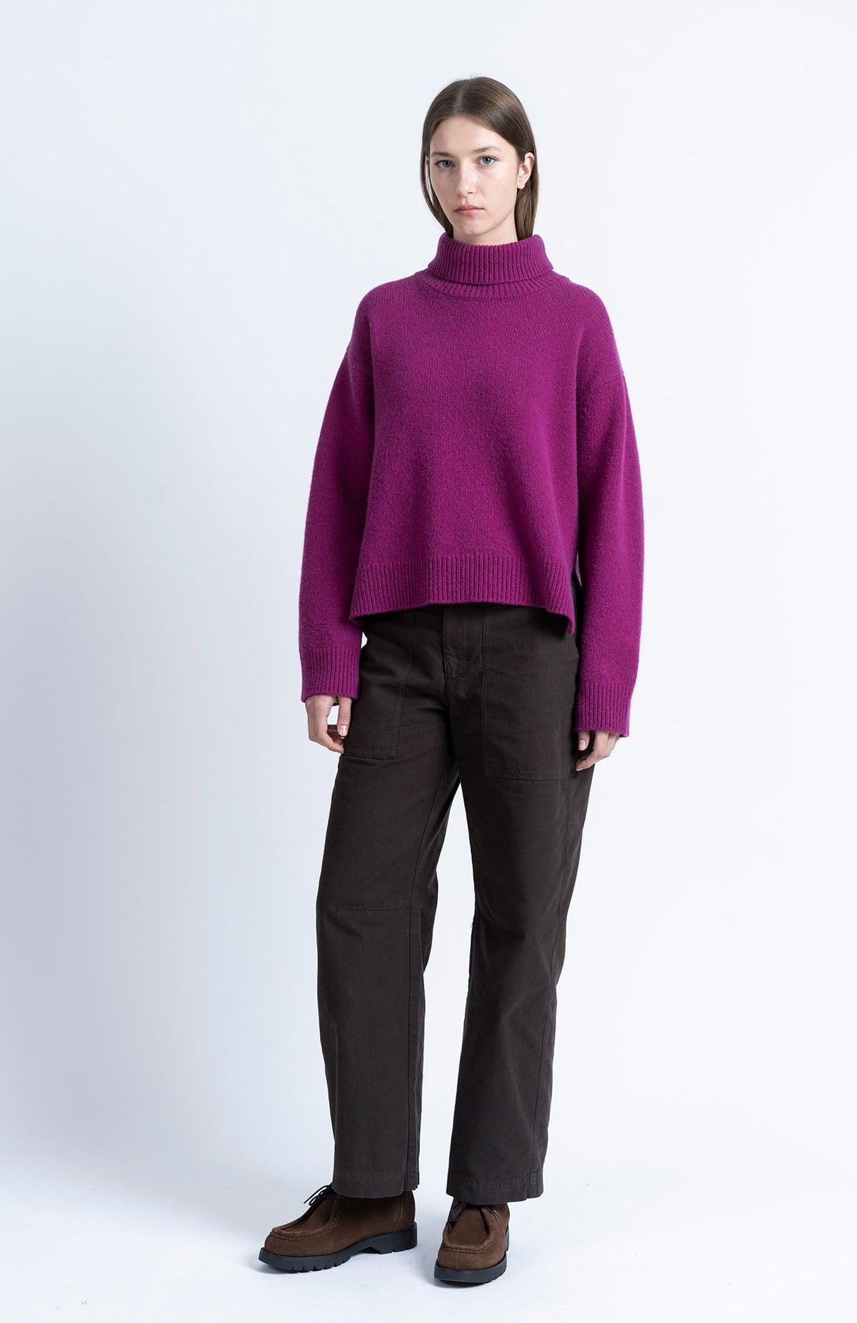 A model stands against a light gray studio background wearing a cropped magenta-purple turtleneck sweater, dark brown straight-leg pants, and brown shoes.