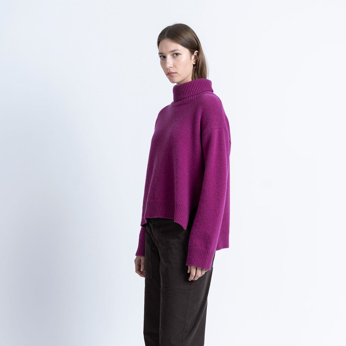 A model stands in profile wearing a bright magenta-purple knit turtleneck sweater, dark brown trousers, and brown leather shoes against a plain white studio background.