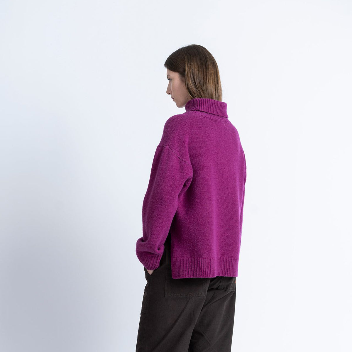 A model is shown from the back wearing a bright magenta-purple knit turtleneck sweater, dark brown wide-leg trousers, and brown suede boots against a plain white studio background.