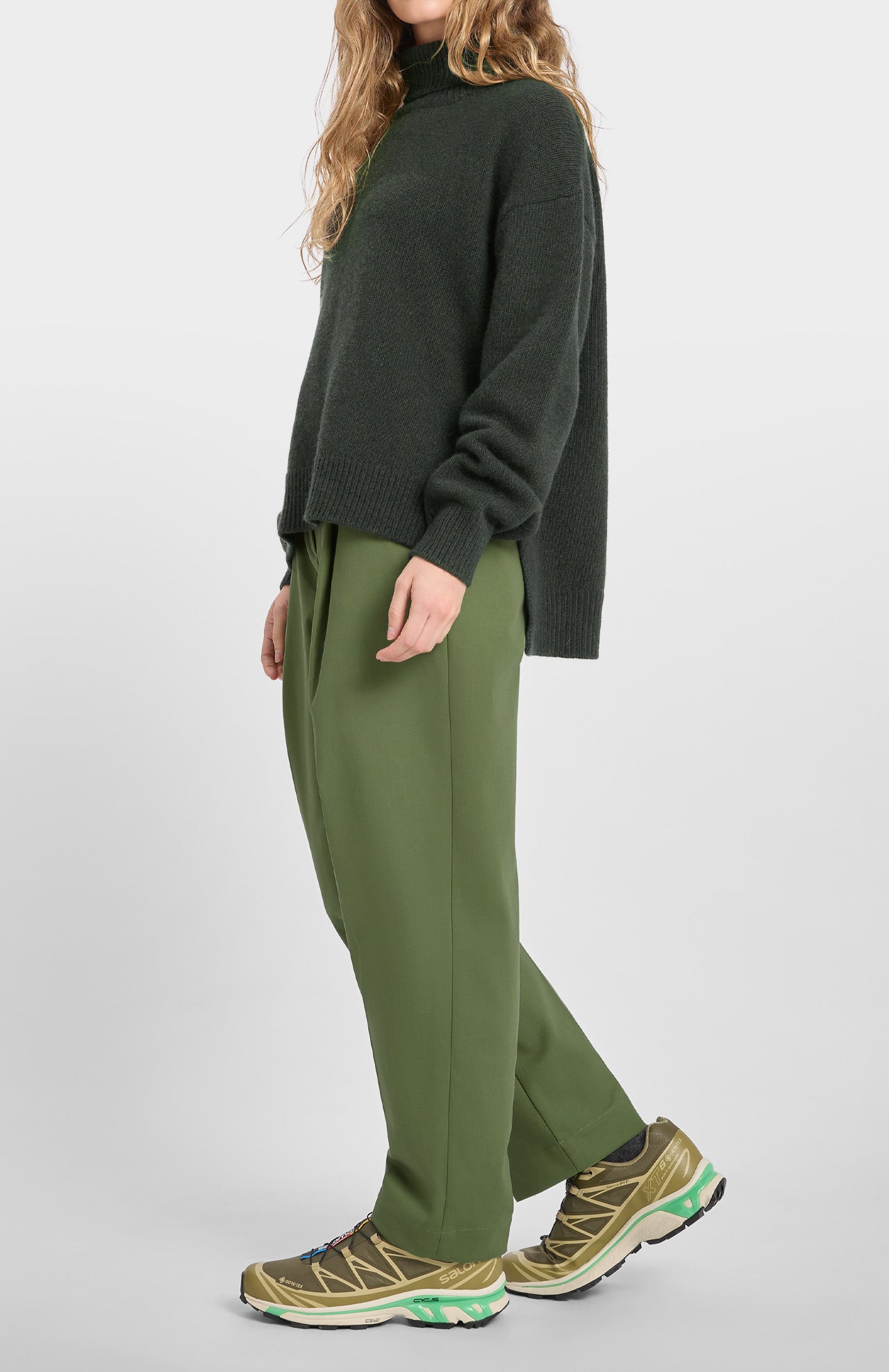 A person wearing the Loreak Mendian Isla W Jumper in forest green, olive green pants, and tan sneakers with green accents stands against a plain white background.