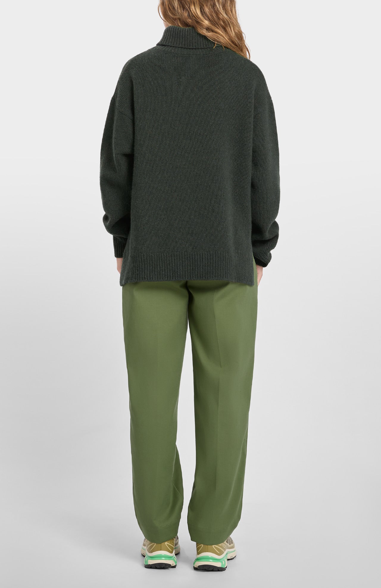 A person stands facing away in a Loreak Mendian Isla W Jumper - Forest Green, paired with light green pants and yellow-green sneakers, set against a plain light background.