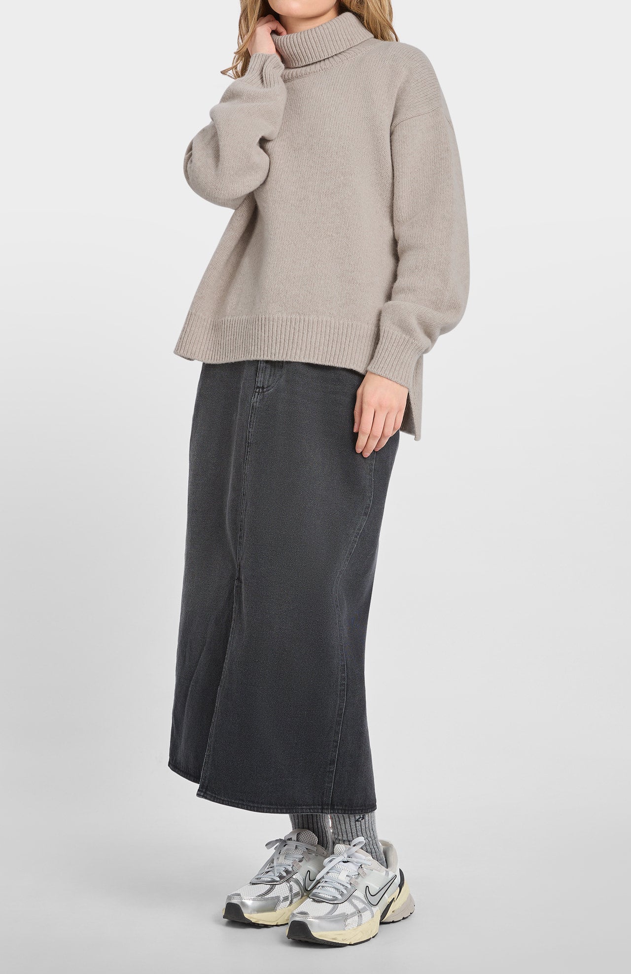 A person stands against a plain background wearing the Loreak Mendian Isla W jumper in cool beige, paired with a long dark denim skirt, gray socks, and metallic silver sneakers.