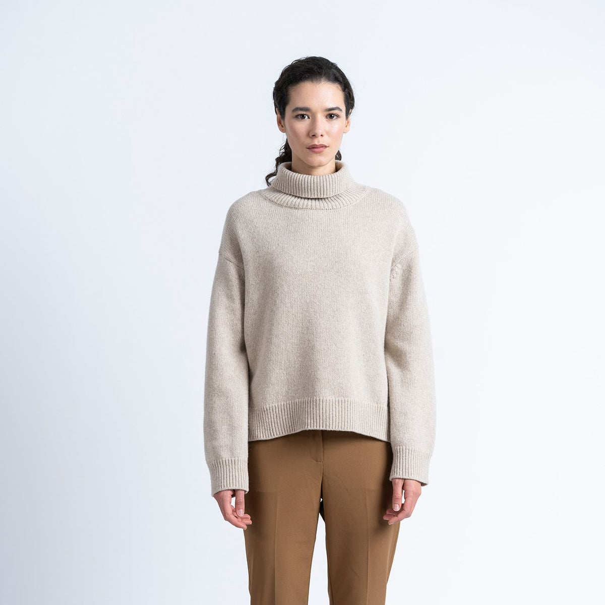 model wears loreak mendian natural colour roll neck knit with camel trousers on a white background.