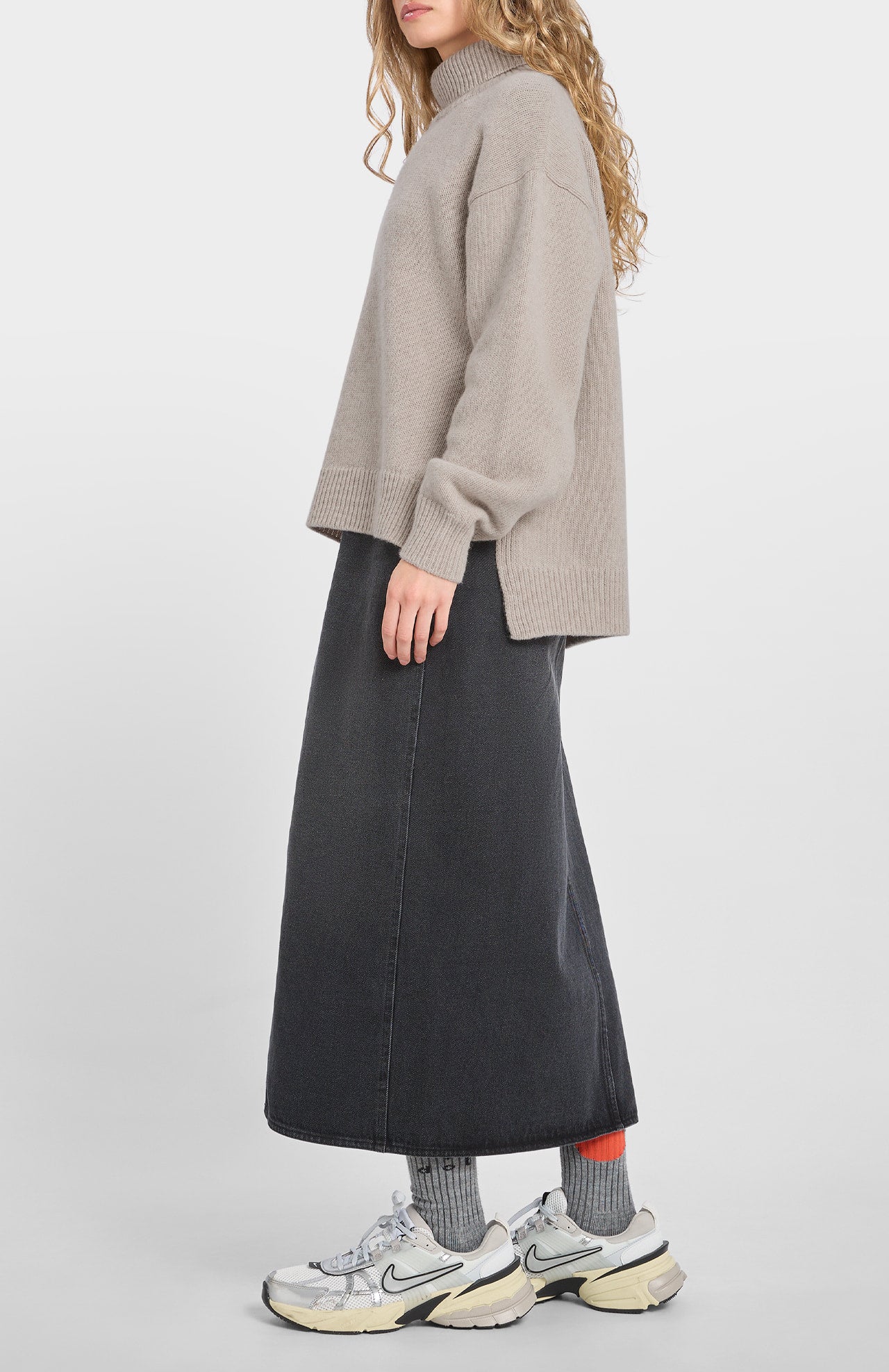 A person wearing the Loreak Mendian Isla W jumper in Cool Beige, a long black denim skirt, gray socks, and white sneakers stands against a plain background.