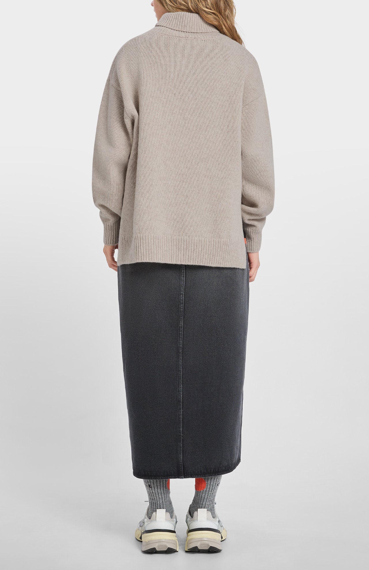 Person standing with their back to the camera, wearing the Loreak Mendian Isla W jumper in Cool Beige, a long black skirt, gray socks, and white sneakers.