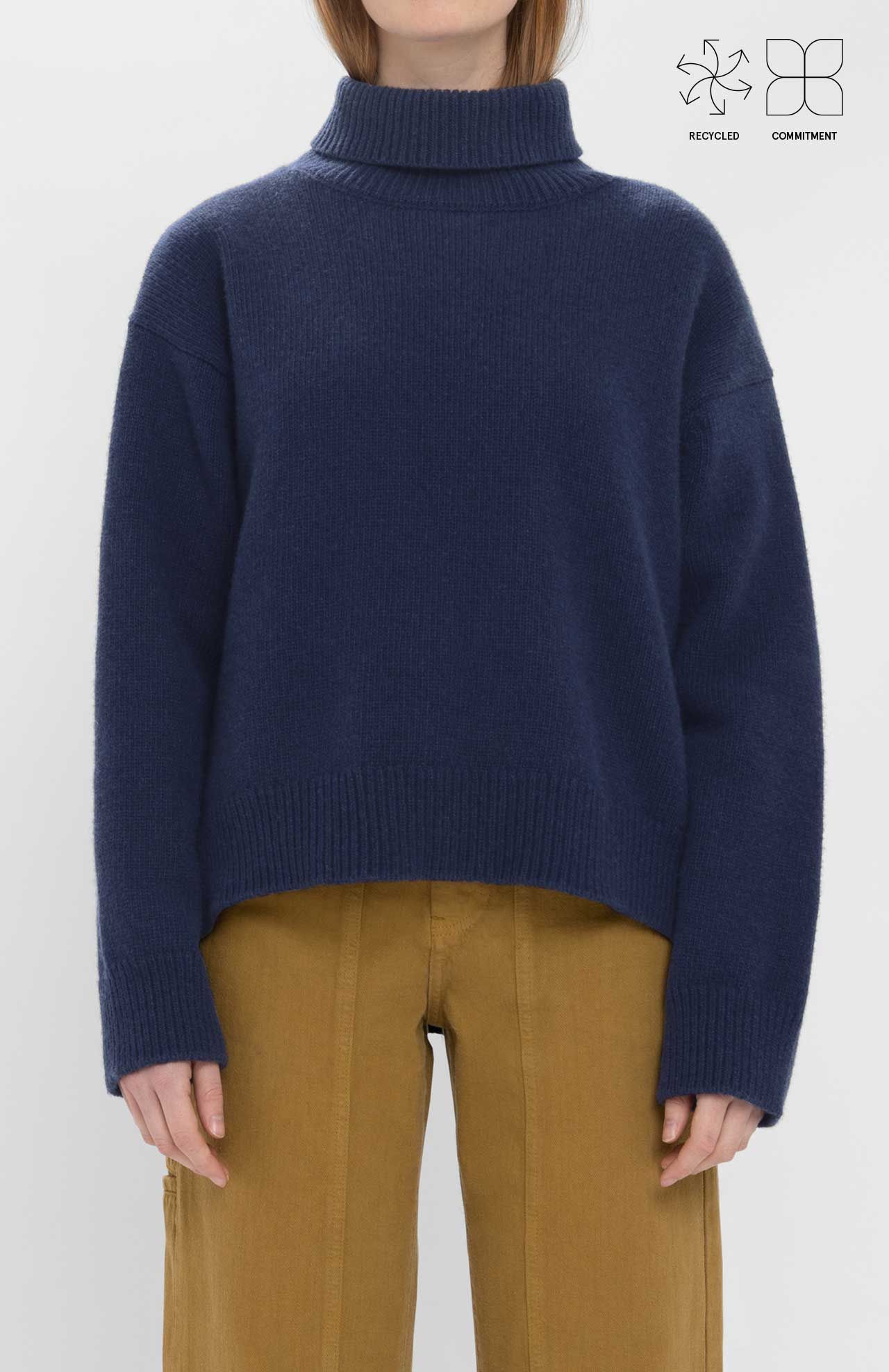 A person wears the Loreak Mendian Isla W jumper, a navy blue chunky knit turtleneck, paired with mustard yellow pants against a white background. Recycled and eco-friendly icons in the corner highlight this sustainable Loreak Mendian sweater.