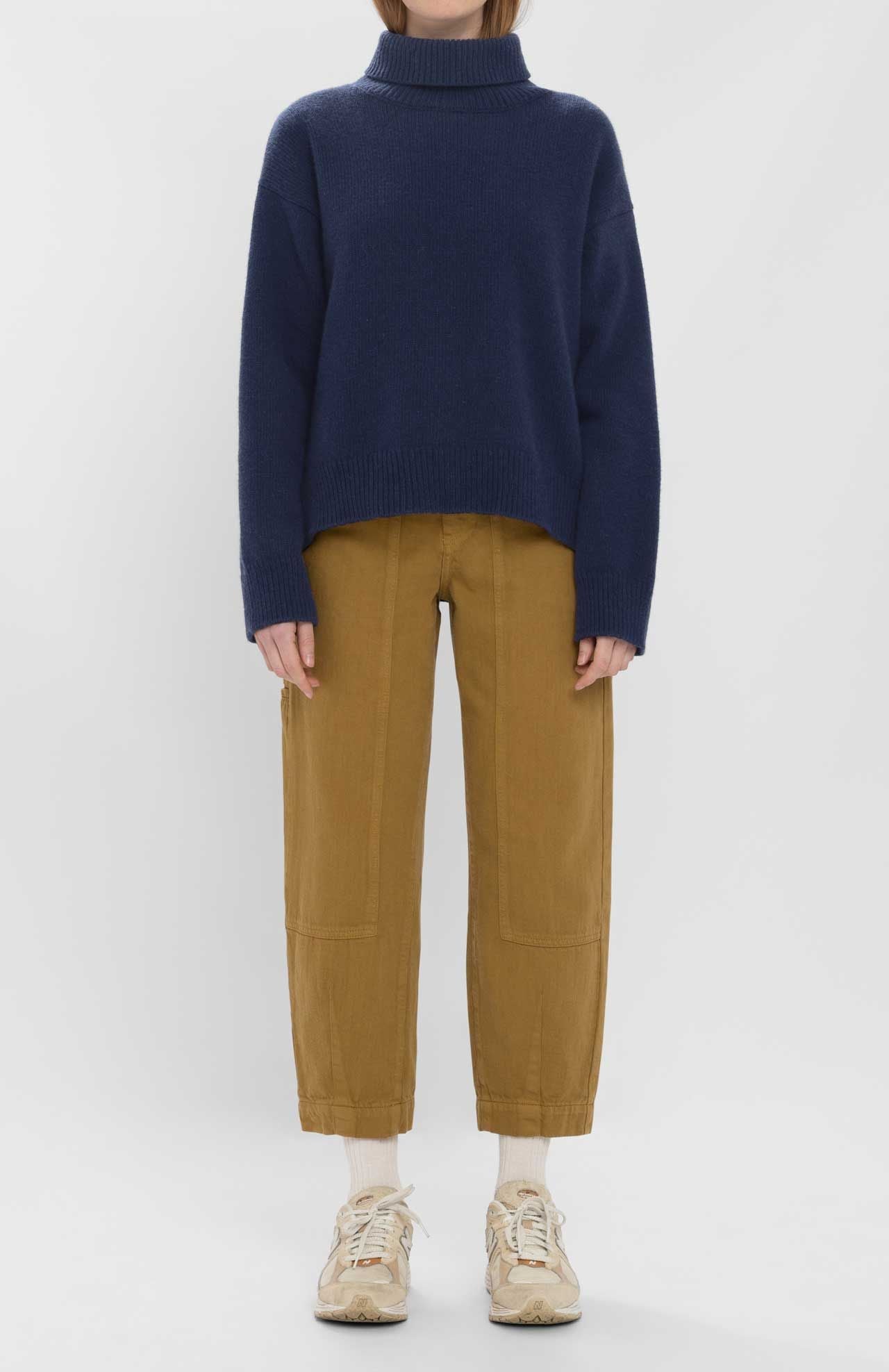 A person stands against a plain background, wearing the Loreak Mendian Isla W jumper—a navy blue chunky knit turtleneck sweater—paired with tan pants, white socks, and beige sneakers.