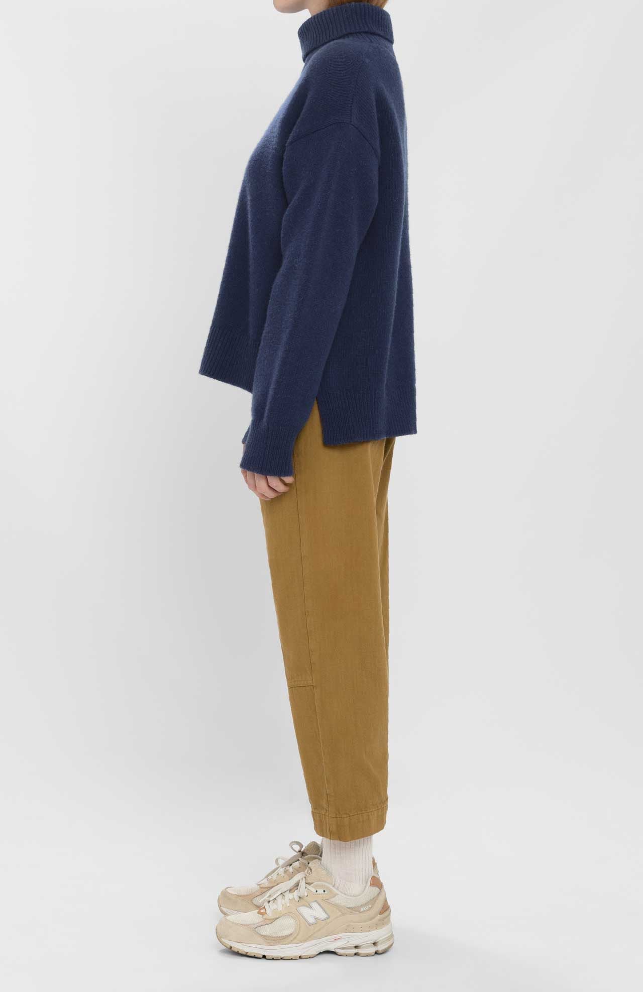 A person in profile wears the Loreak Mendian Isla W jumper—a navy chunky knit turtleneck—paired with tan pants, cream socks, and beige sneakers against a plain white background.