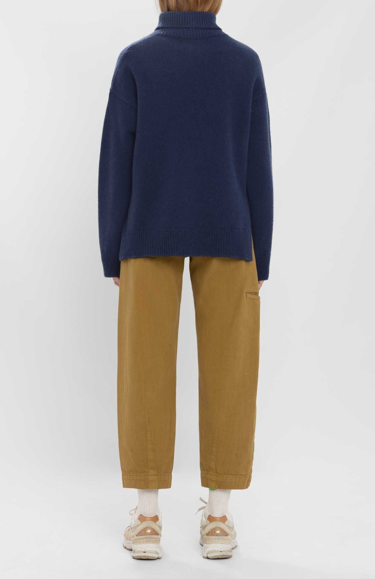 A person stands facing away, wearing the Loreak Mendian Isla W jumper by Loreak Mendian in navy, paired with tan pants, white socks, and beige sneakers against a plain light background.