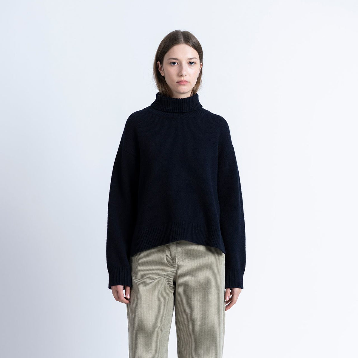 a woman wears navy rollneck knitted sweater with beige coloured cords on a white background