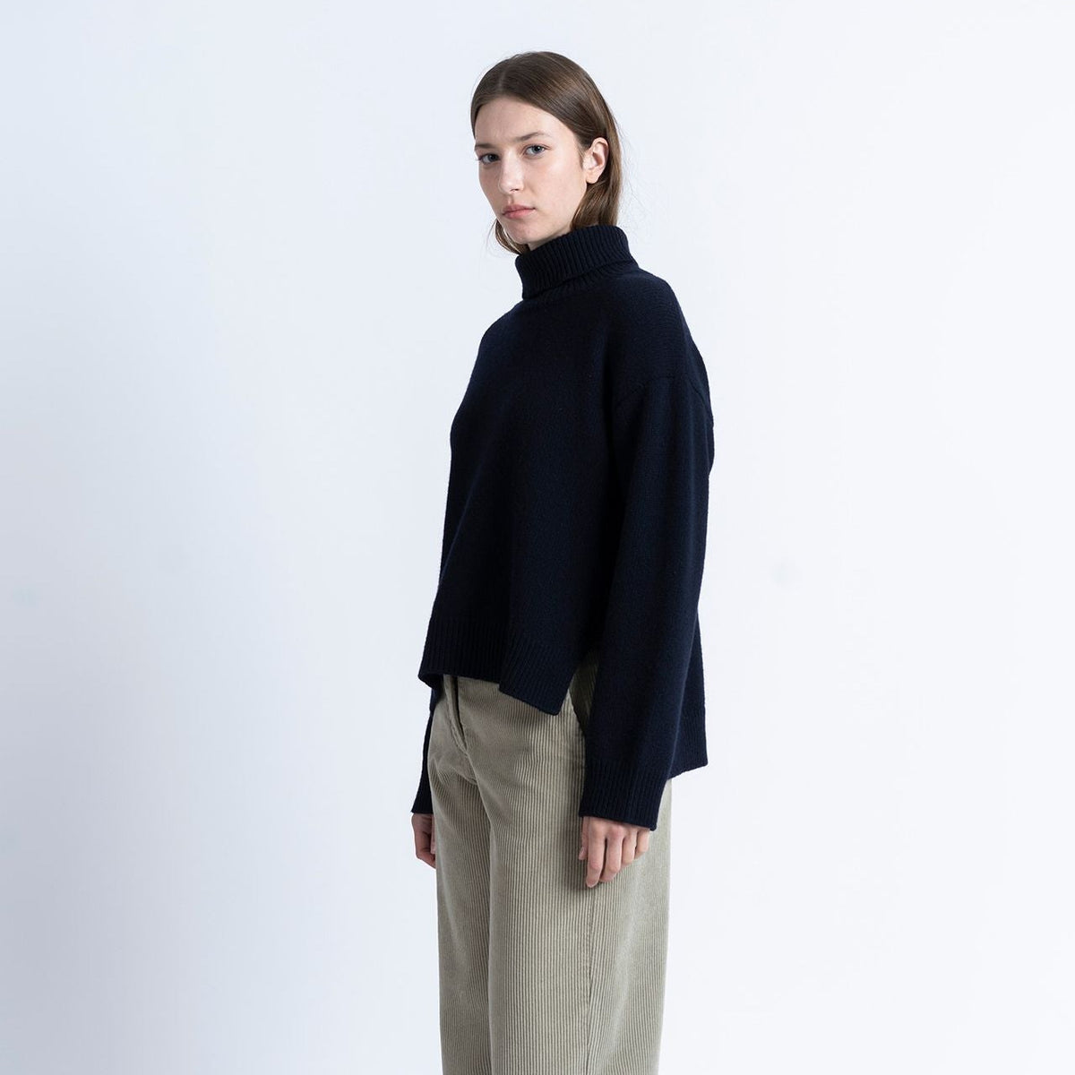A model stands in profile wearing a dark navy turtleneck sweater, beige wide-leg trousers, and brown boat shoes against a plain white studio background.