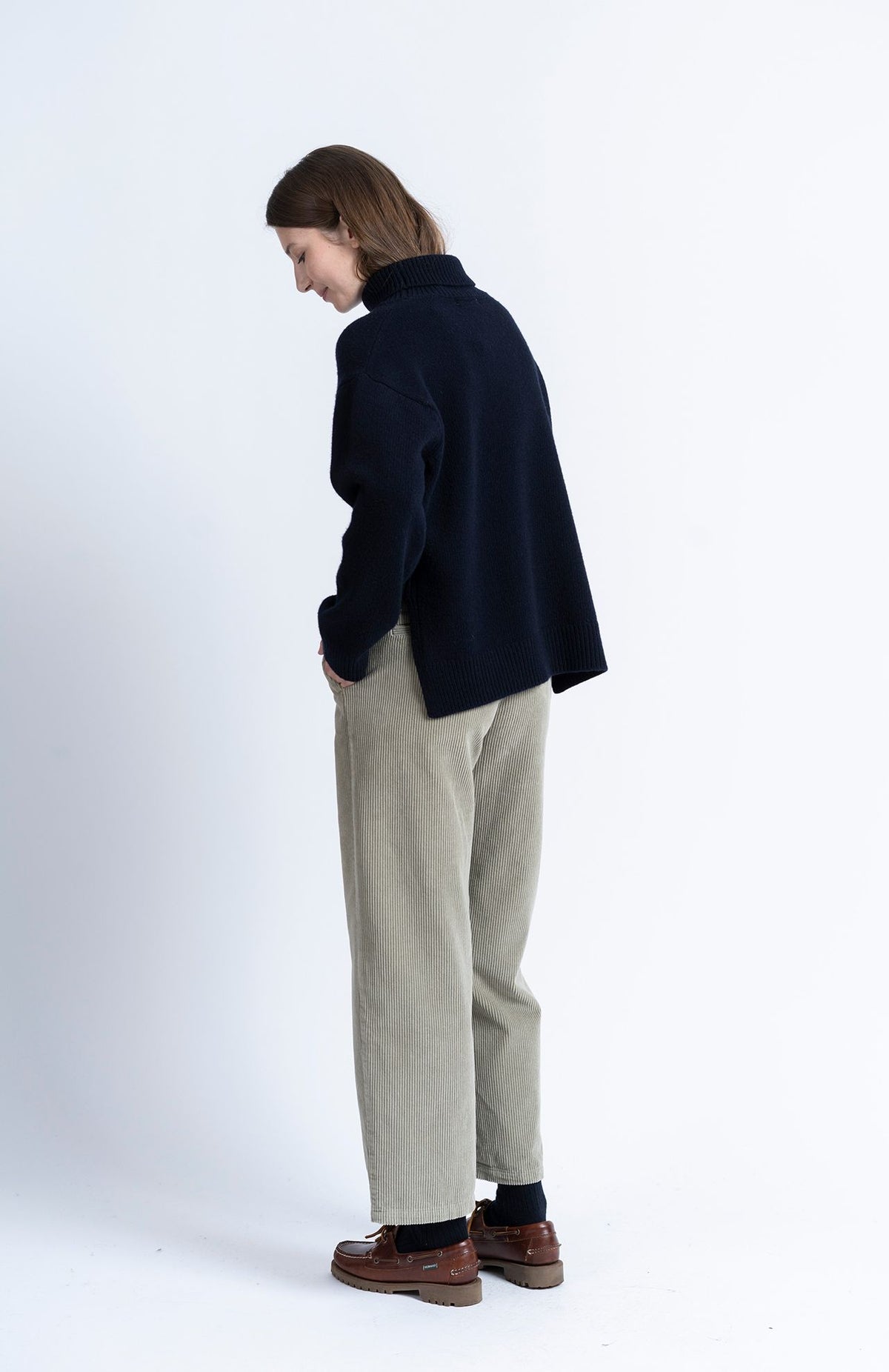 A model wearing a dark navy blue turtleneck sweater, light beige corduroy trousers, and brown leather shoes, standing in profile against a plain white studio background.