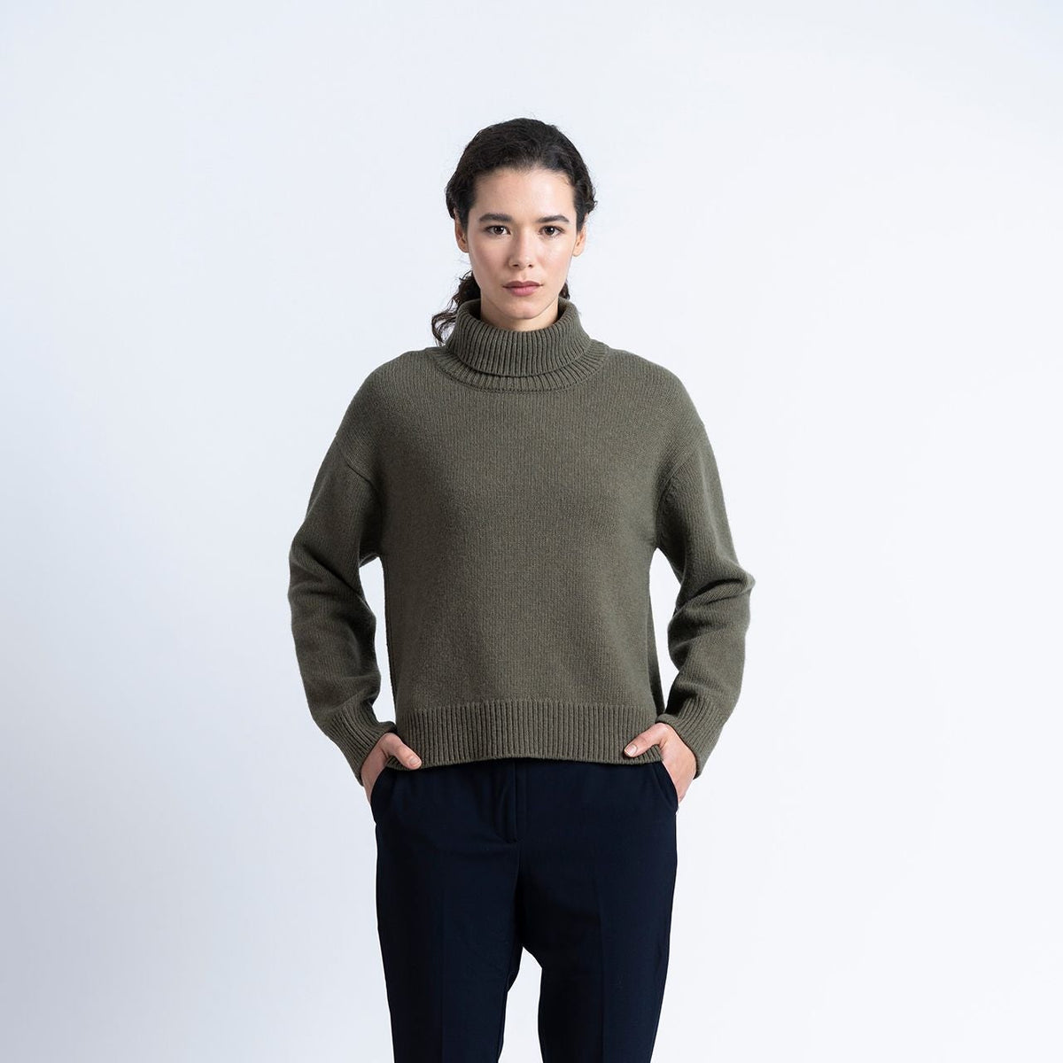 a woman wears a khaki green rollneck wool knit and navy trousers on a white background
