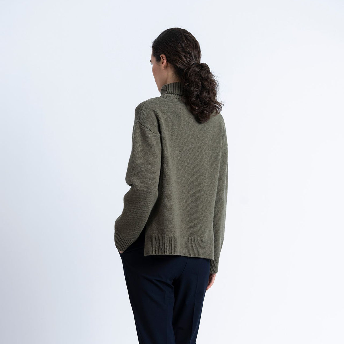 a woman wears a khaki green rollneck wool knit and navy trousers on a white background