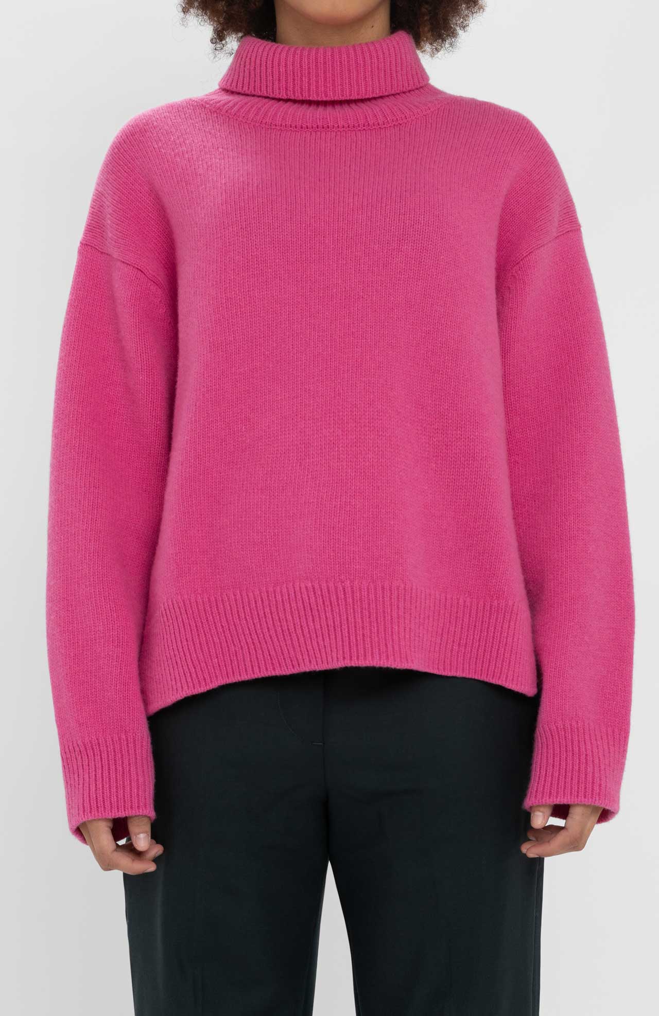 A person wearing a **Loreak Mendian Isla W jumper**, a bright pink, cozy knit turtleneck sweater, and black pants stands against a plain background.