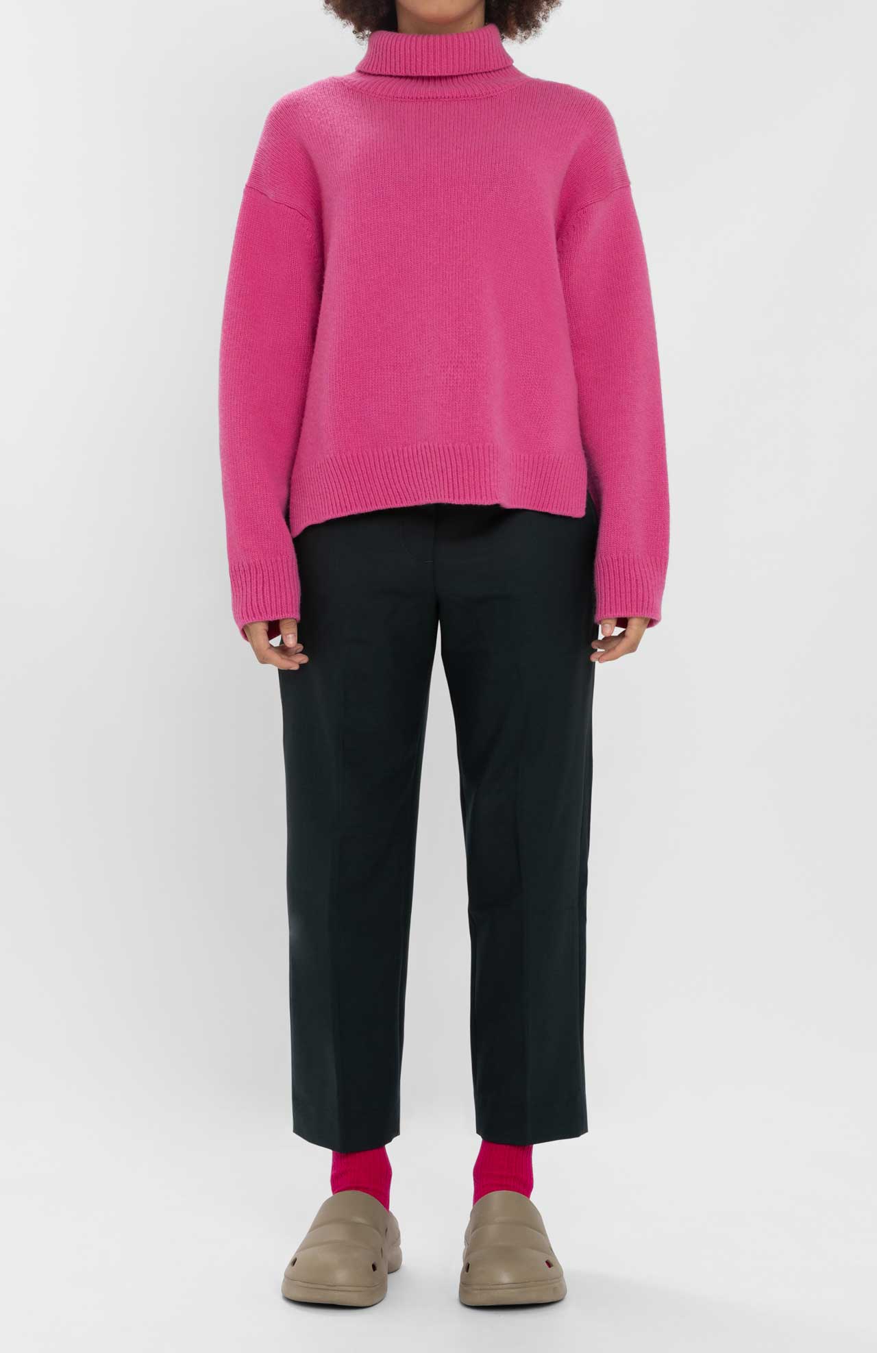 A person standing against a plain white background, wearing the Loreak Mendian Isla W jumper in bright pink, black pants, pink socks, and beige slip-on shoes.