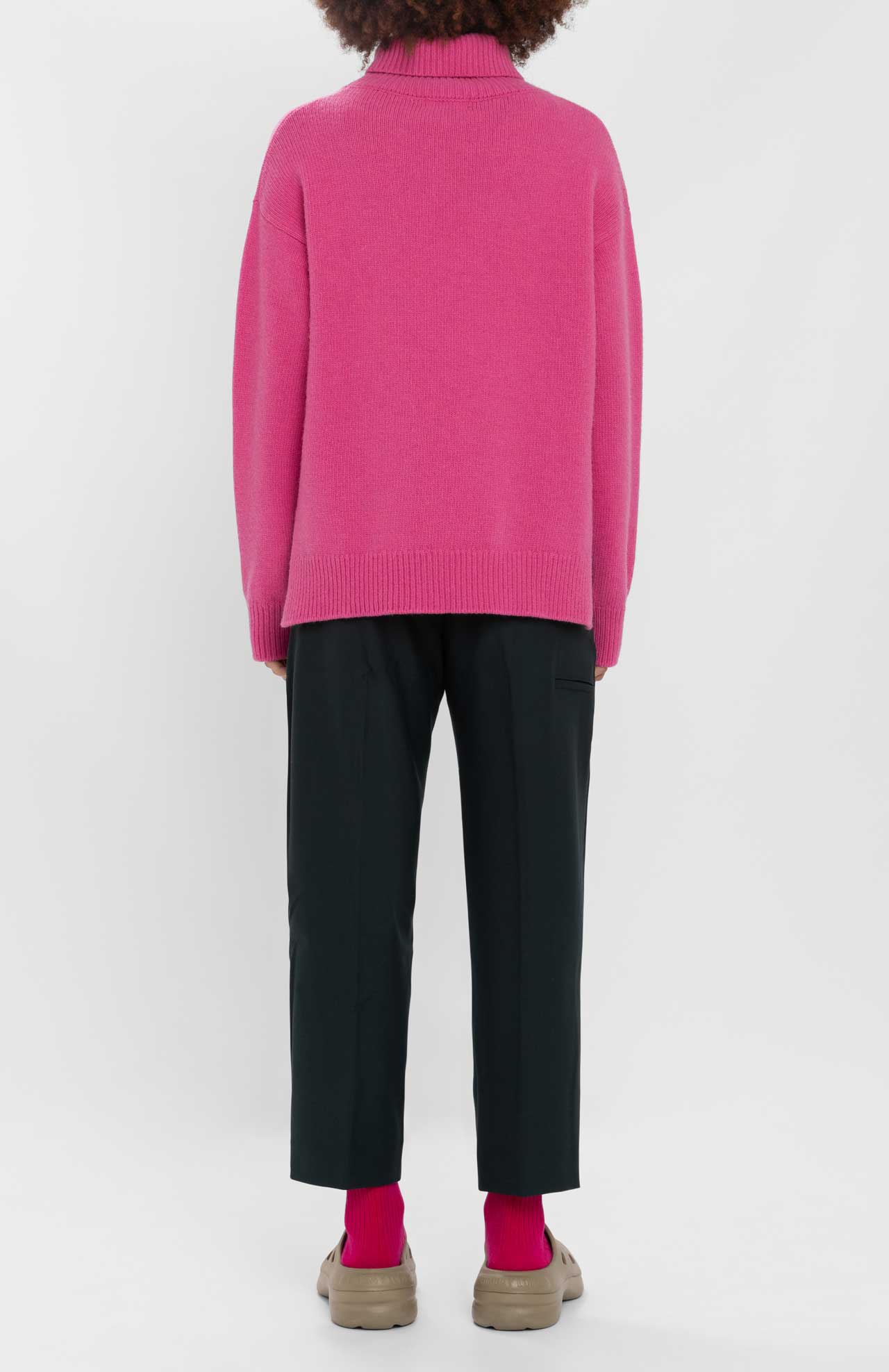 A person is standing with their back to the camera, wearing a bright pink Loreak Mendian Isla W jumper, black pants, and beige shoes.