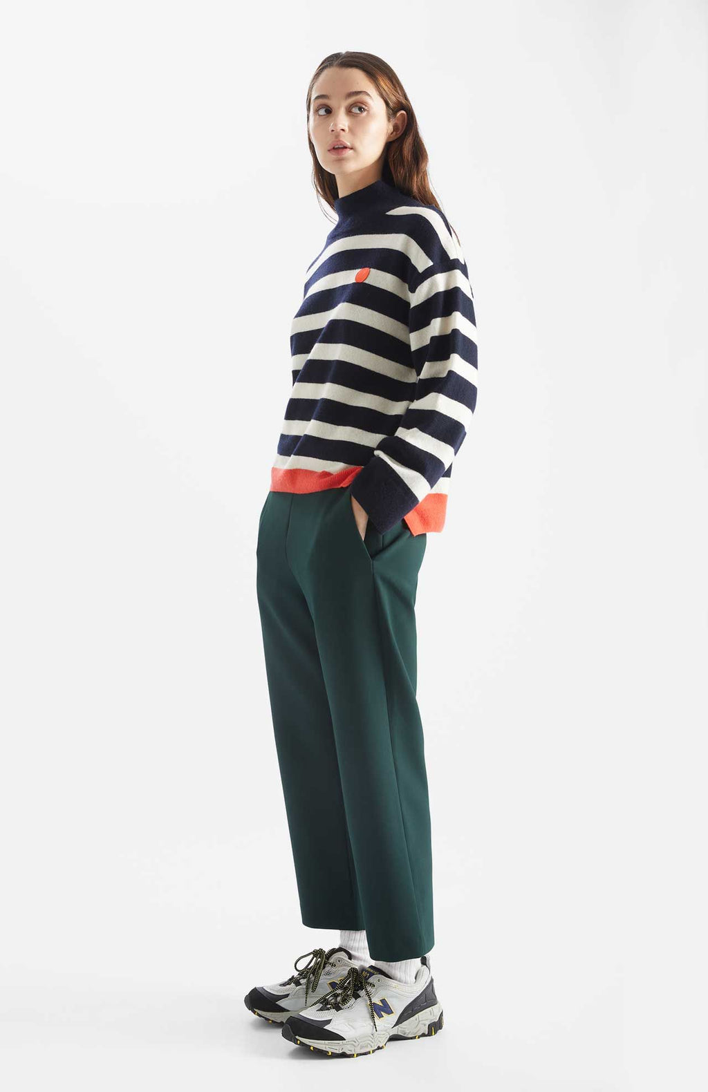 Person wears Kada Dot jumper in a navy and white stripe. There is one songle orange stripe at the base and an orange dot at the chest. Side view.