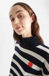 Person wears Kada Dot jumper in a navy and white stripe. There is one songle orange stripe at the base and an orange dot at the chest. Close up of orange dot.