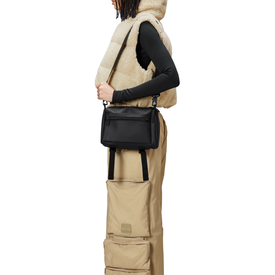 Rains texel crossbody bag blackperson is using the shoulder strap to carry a Matte Black crossbody bag with side strap for suitcase carry