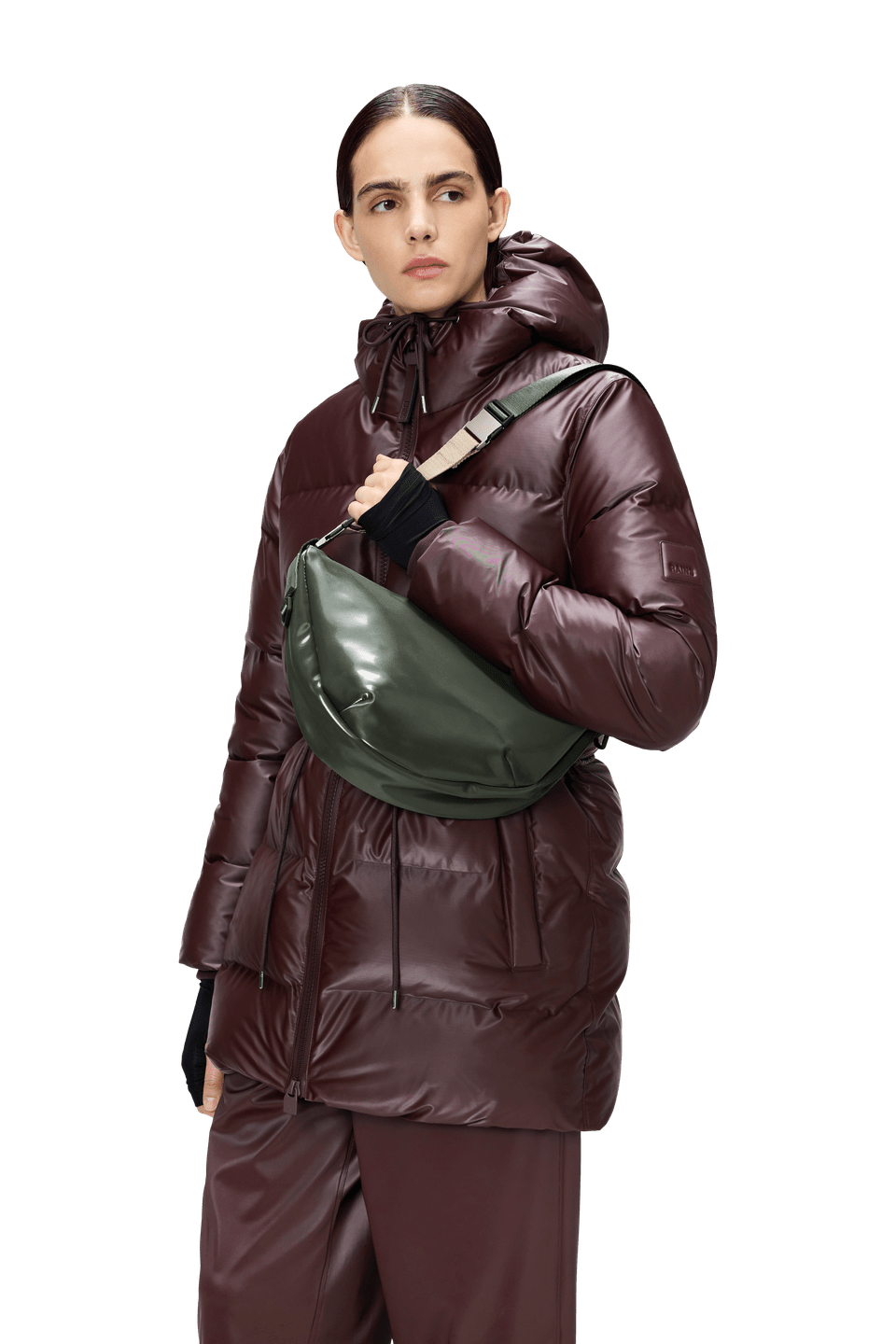 A person in a shiny maroon puffer jacket and pants with the hood up carries a Rains Valera Shoulder Bag in olive green across their body, standing against a plain background.