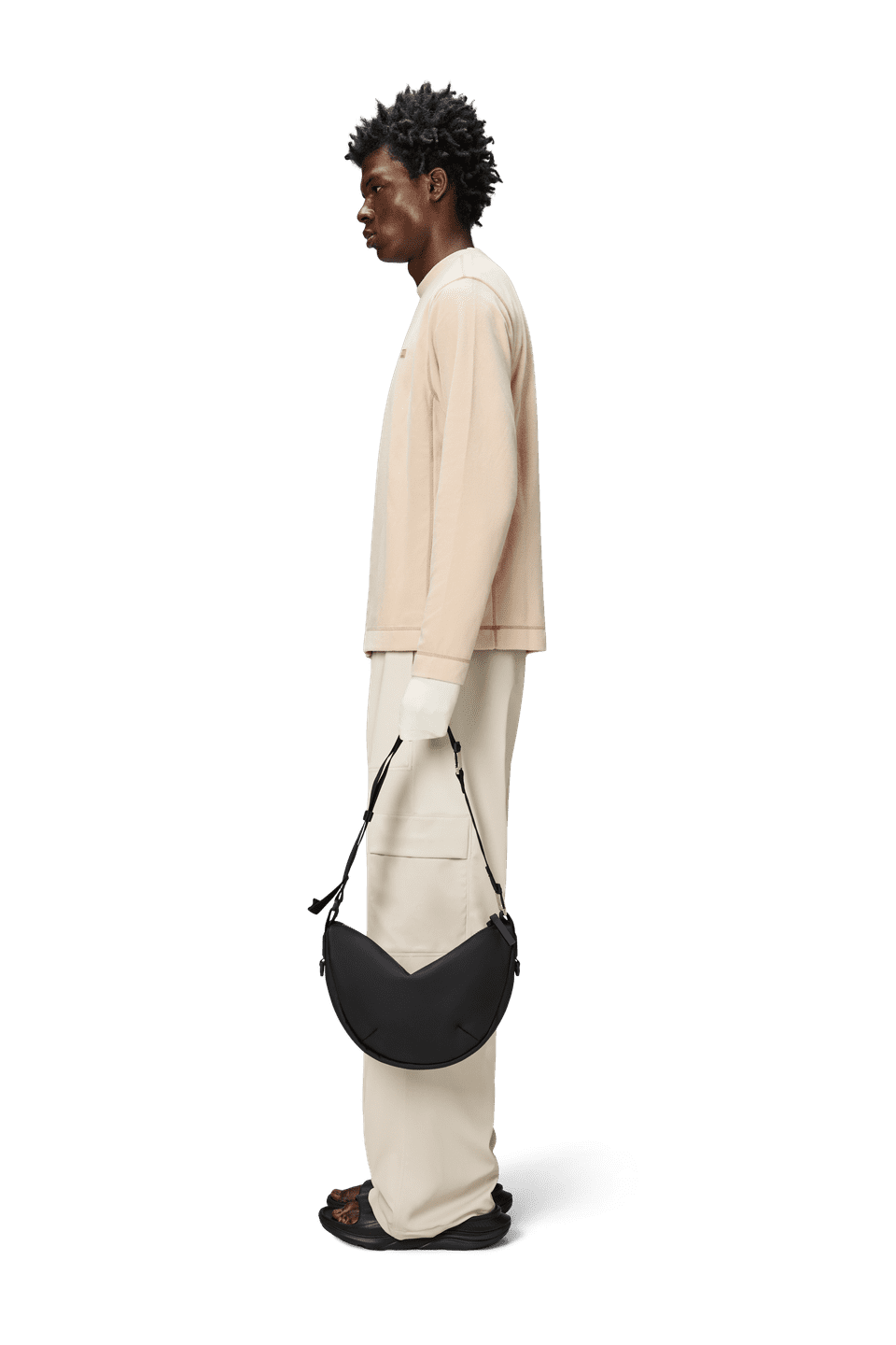 A person in profile wears a beige top and pants, holding a Rains Valera Shoulder Bag with an adjustable strap, crafted from waterproof PU fabric.