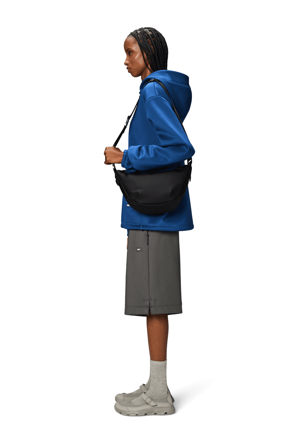 A person in a blue jacket and gray shorts stands sideways, wearing light-colored socks and shoes. They're carrying a sleek Rains Valera Shoulder Bag made from waterproof PU fabric, featuring an adjustable strap for comfort.