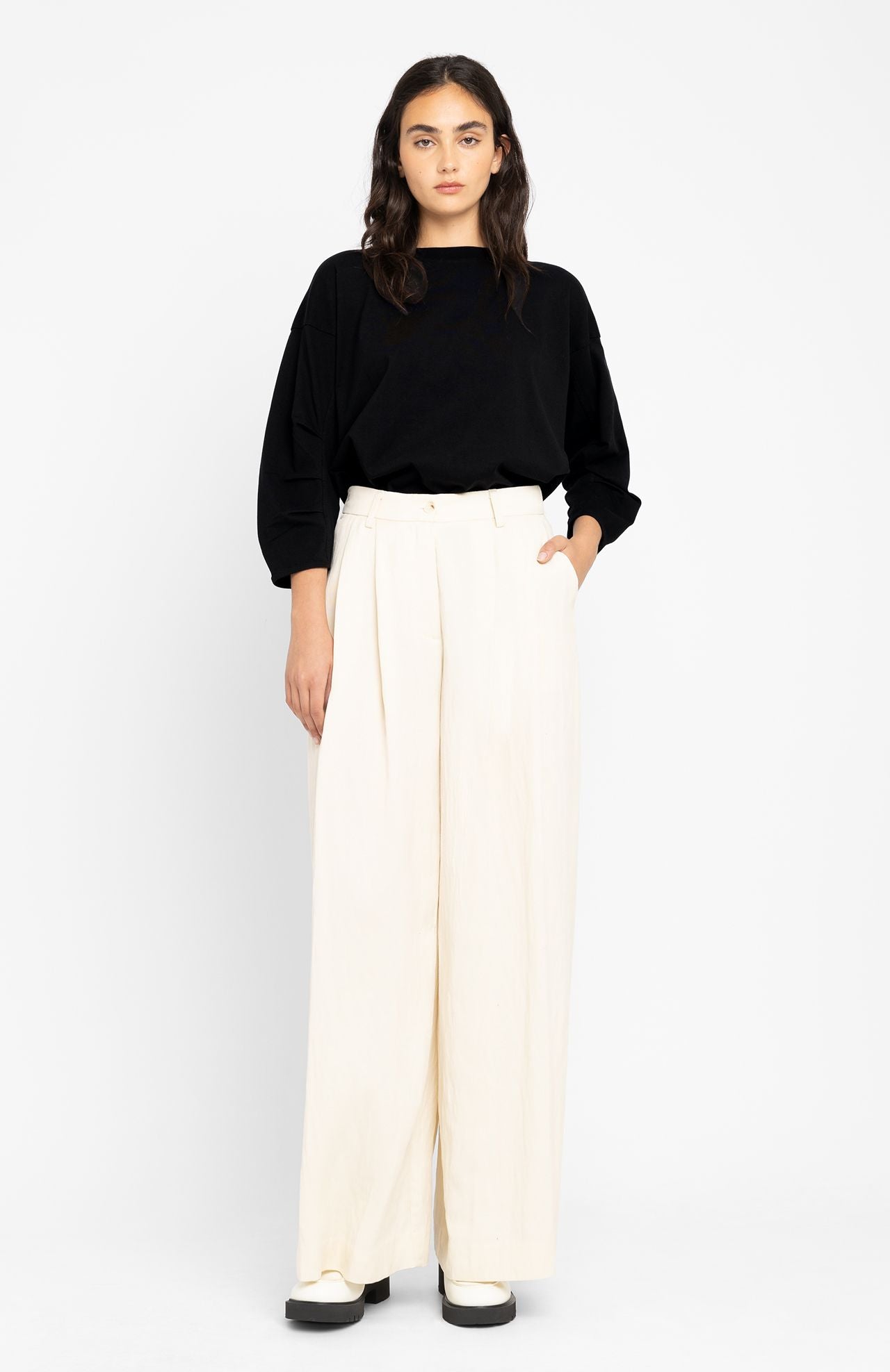 A person with long hair stands against a white backdrop, wearing the Loreak Mendian Zuri Blouse in black, characterized by its oversized fit and pleated sleeves, paired with white wide-legged pants.