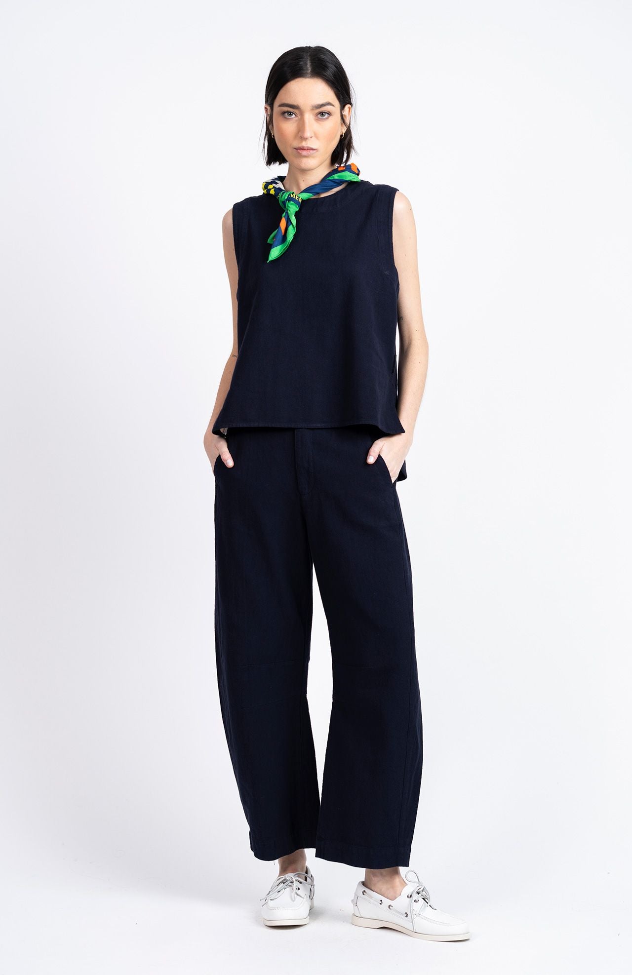 A woman stands against a white background wearing Loreak Mendian's Bera Sleeveless Top in navy, paired with wide-leg pants, white sneakers, and a green and yellow scarf around her neck, hands in pockets.