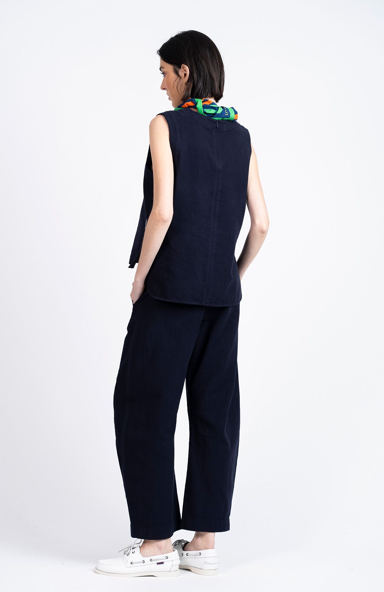 A person stands facing away, wearing the Loreak Mendian Bera Sleeveless Top in navy, matching wide-leg pants, white sneakers, and a colorful scarf around their neck.