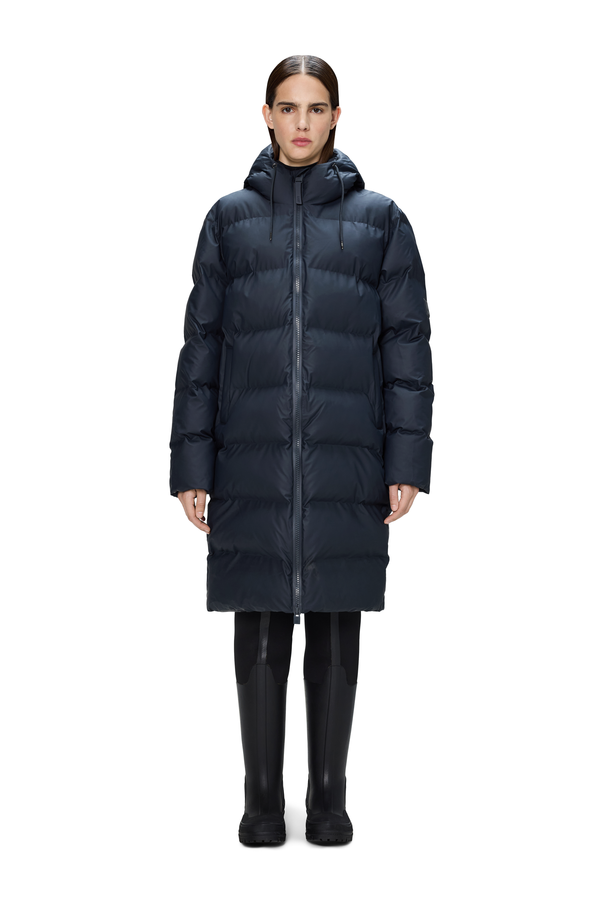 A person wears the Rains Alta Puffer Longer in navy, featuring featherless insulation, paired with black pants and boots, standing facing forward against a plain white background.