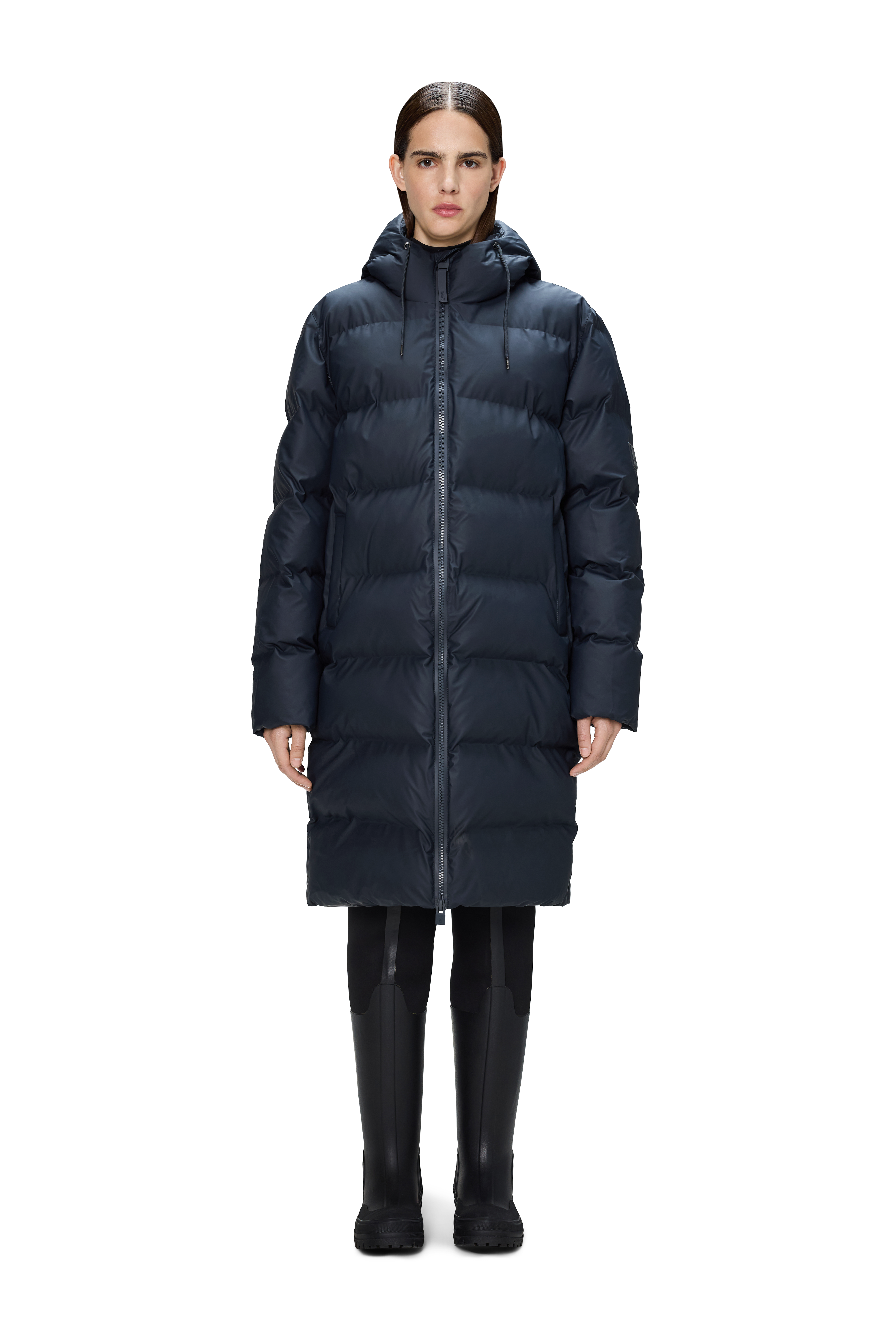 A person wears the Rains Alta Puffer Longer in navy, featuring featherless insulation, paired with black pants and boots, standing facing forward against a plain white background.