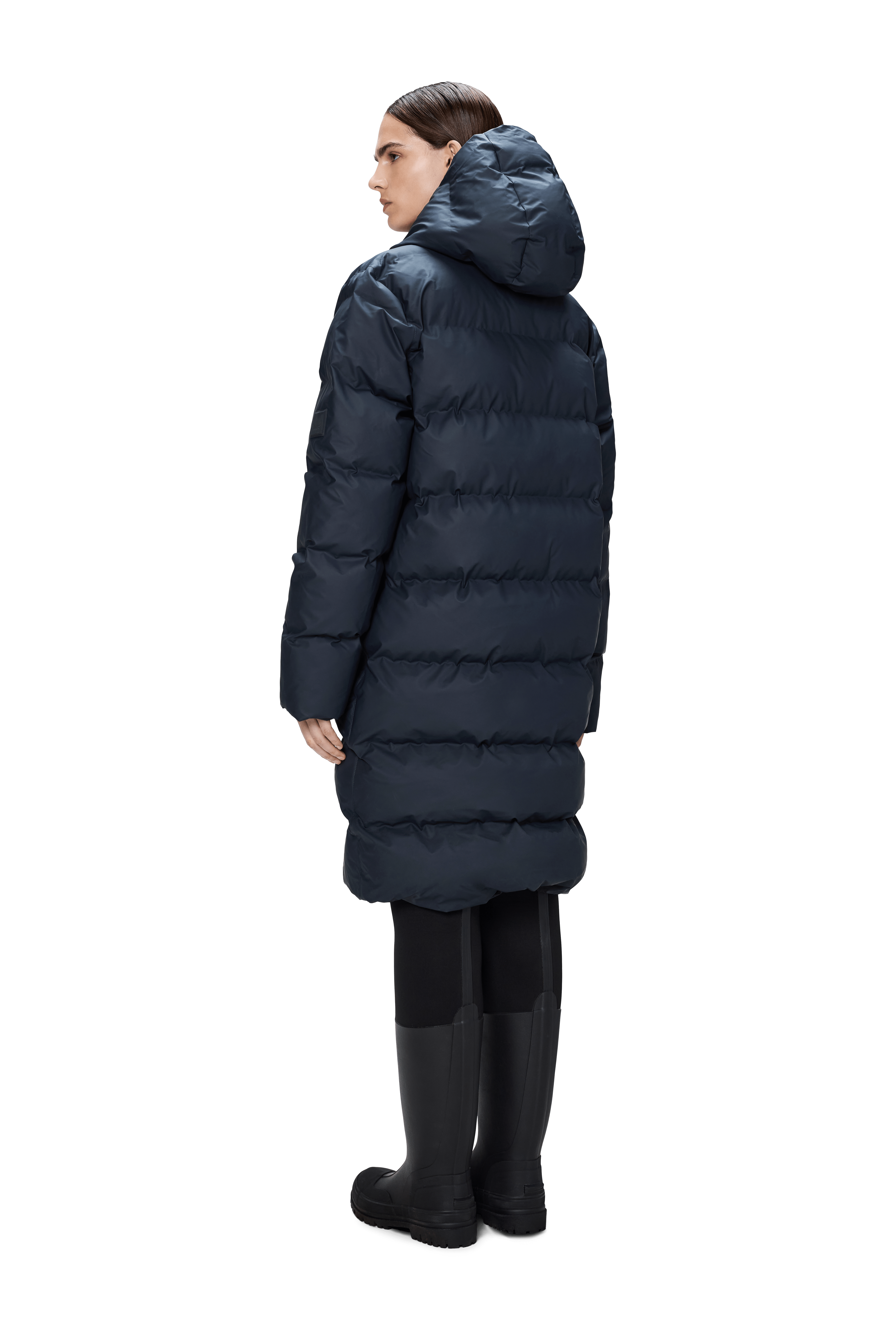 A person models the Rains Alta Puffer Longer in Navy, a featherless, waterproof jacket, paired with black leggings and tall black boots against a plain white background, facing slightly away.