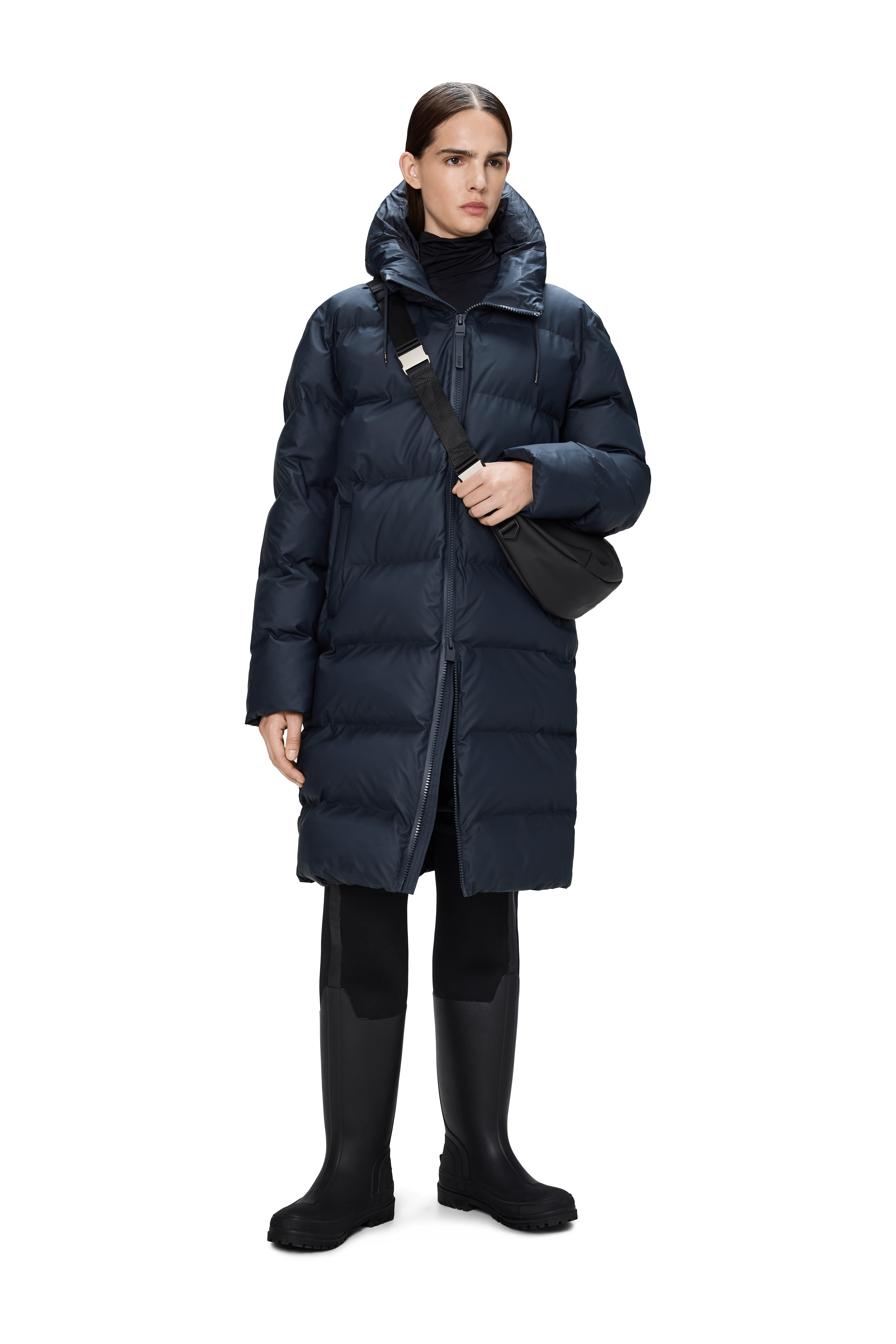 A person wears the Rains Alta Puffer Longer in navy, featuring featherless insulation, black pants, rubber boots, and a black crossbody bag, standing against a plain white background.